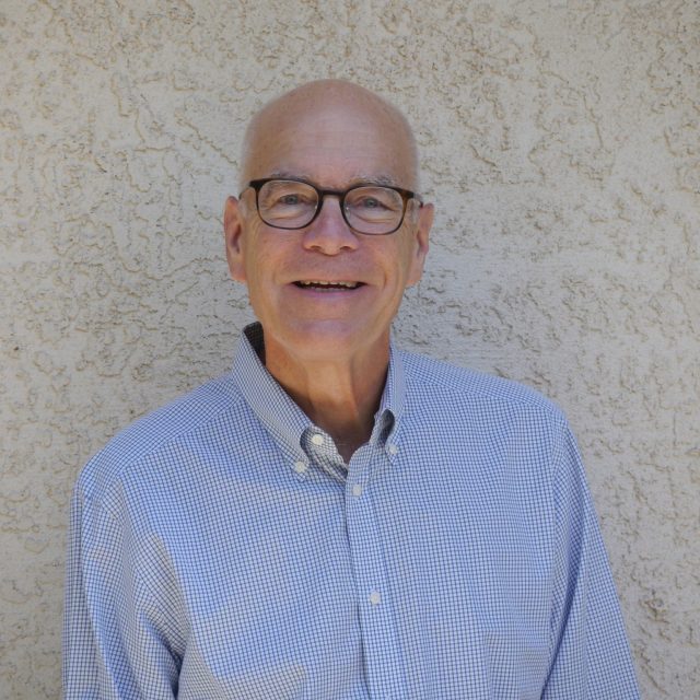 Chuck Champlin - bald, white, male-presenting person with black-framed glasses and blue button-down shirt in front of stucco wall