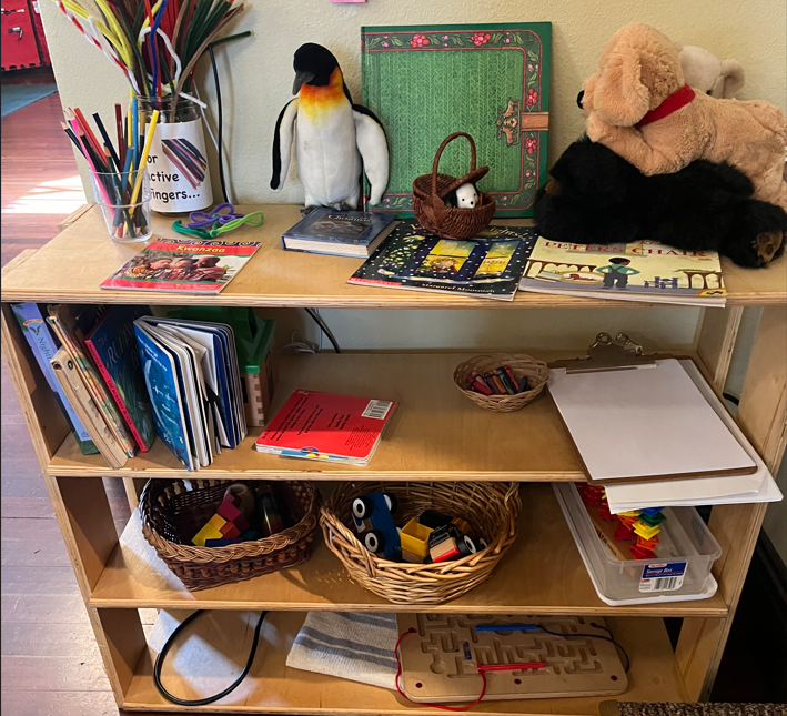 shelf in UUCM sanctuary with stuffed animals, books, pipe cleaners, clipboards with paper, small figurines, and various toys