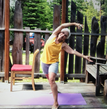 Bettina, a light-skinned female-presenting person with short white hair wearing dark glasses, yellow tank top and grey shorts bends her arms over her head and leans her torso to the right with legs spread in warrier-type pose on a wooden deck
