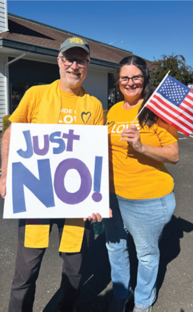 Rev. Kevin Tarsa holds a sign that says, "Just NO!" standing with Rochelle Davisson, who is holding a small USA flag