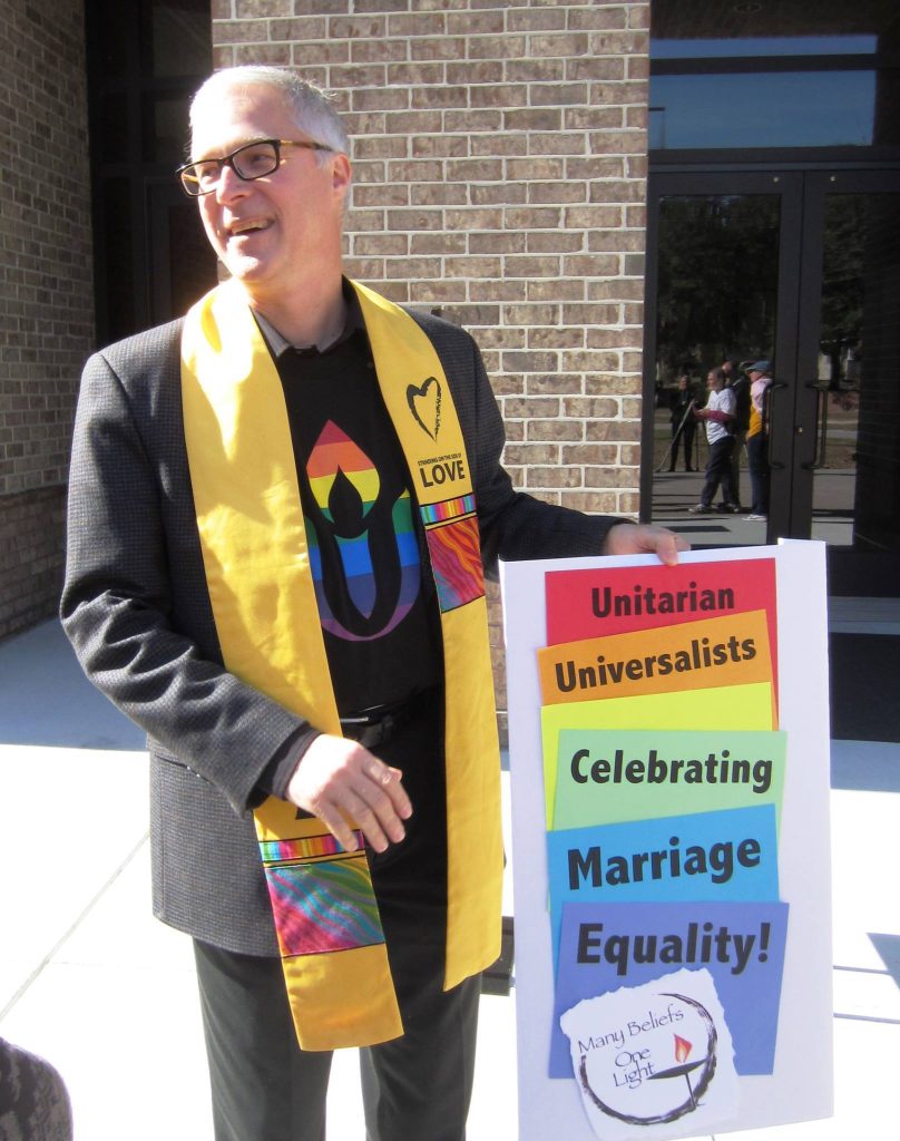 Rev. Kevin in black suit jacket over a rainbow-colored UUA logo t-shirt and yellow "Side With Love" stoll, holding a rainbow-colored sign that reads, "Unitarian Universalists Celebrating Marriage Equality!"