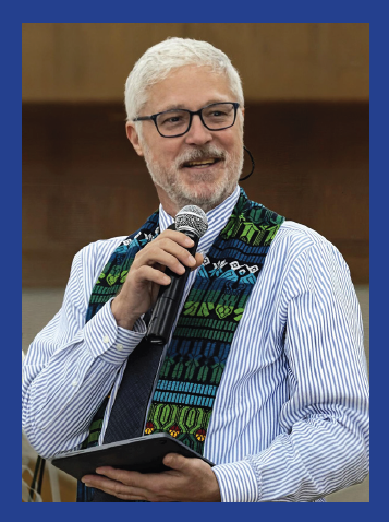 Rev. Kevin Tarsa, with black-framed glasses, holding a microphone, smiling, wearing an intricately embroidered stoll and blue + white pinstripe button down